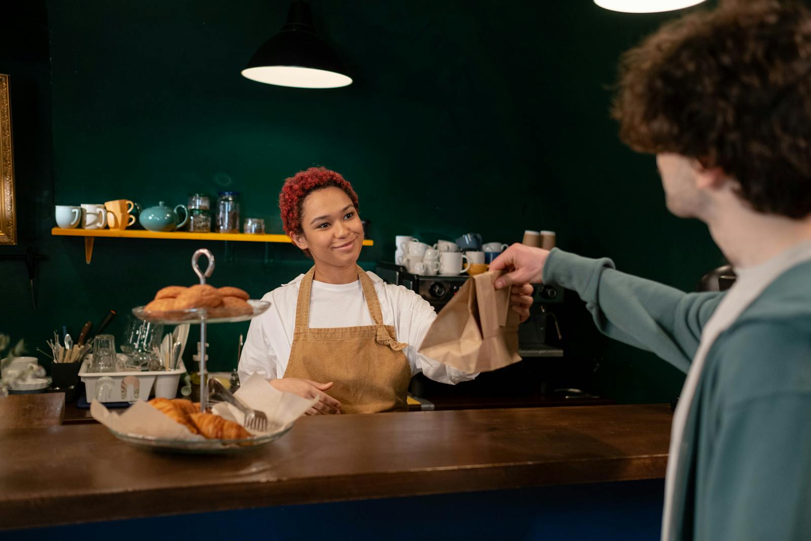 Bäckerei-Verkäuferin reicht freundlich eine Papiertüte mit Backwaren über die Theke an einen Stammkunden.