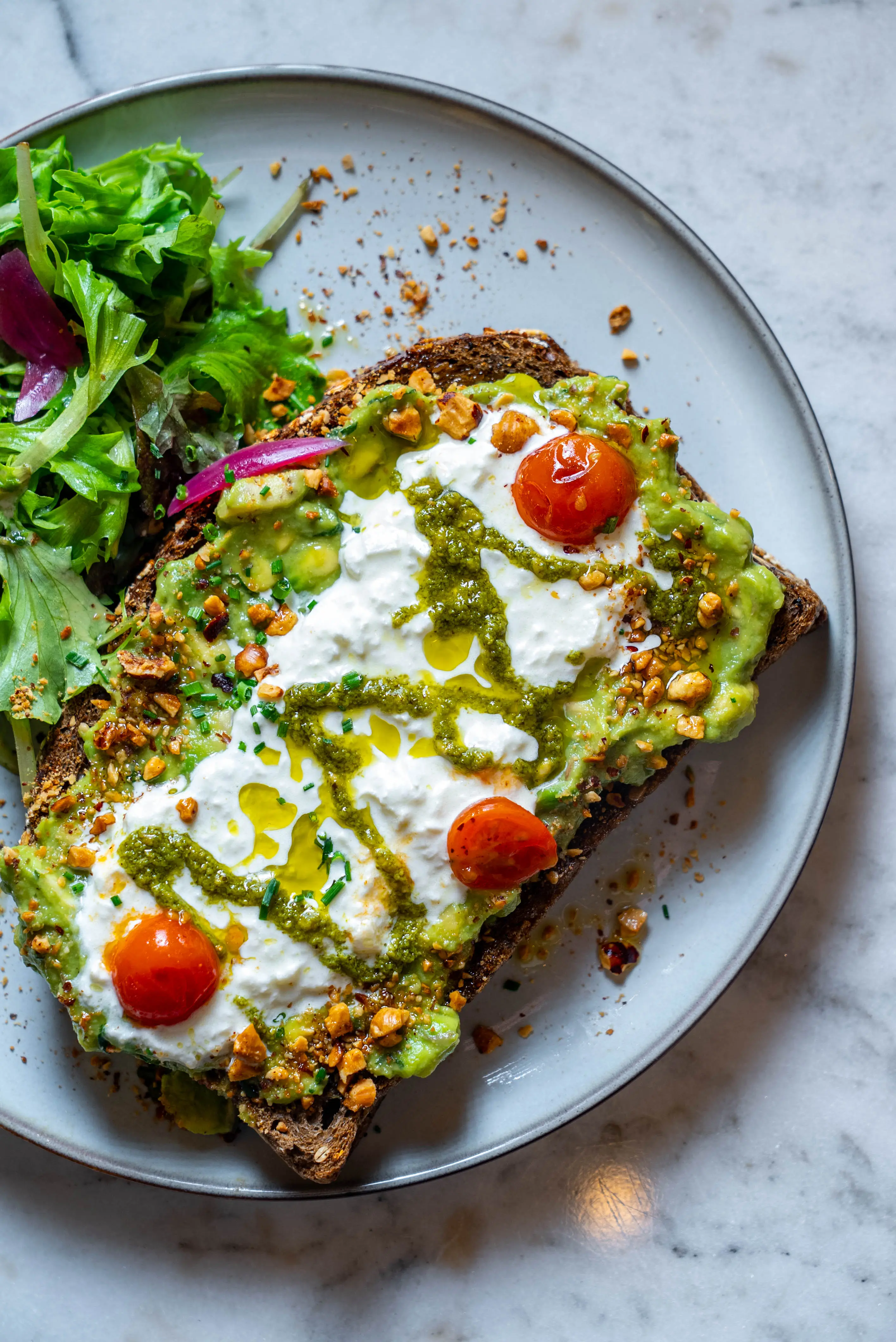 Avocado-Brot mit Pesto, Mozzarella und Kirschtomaten auf hellem Marmor, klar inszeniert.