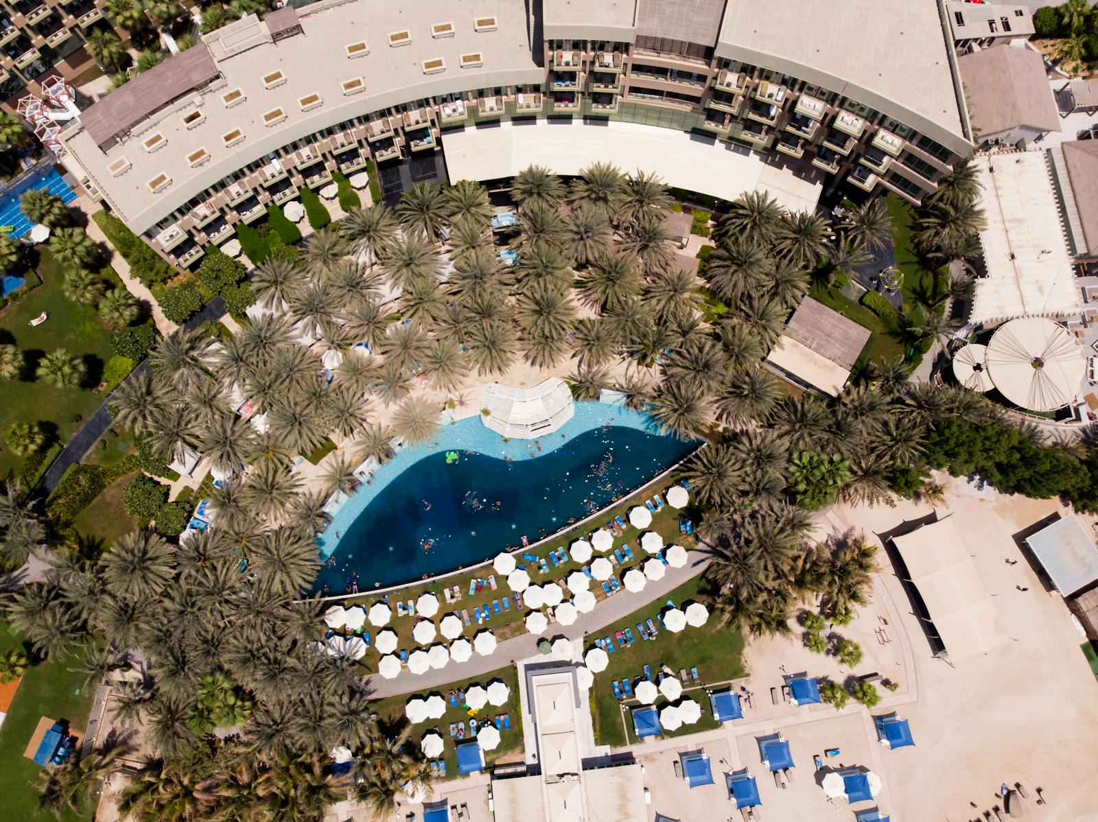 Hotelanlage mit großem Pool, Liegestühlen und Palmen aus der Vogelperspektive.