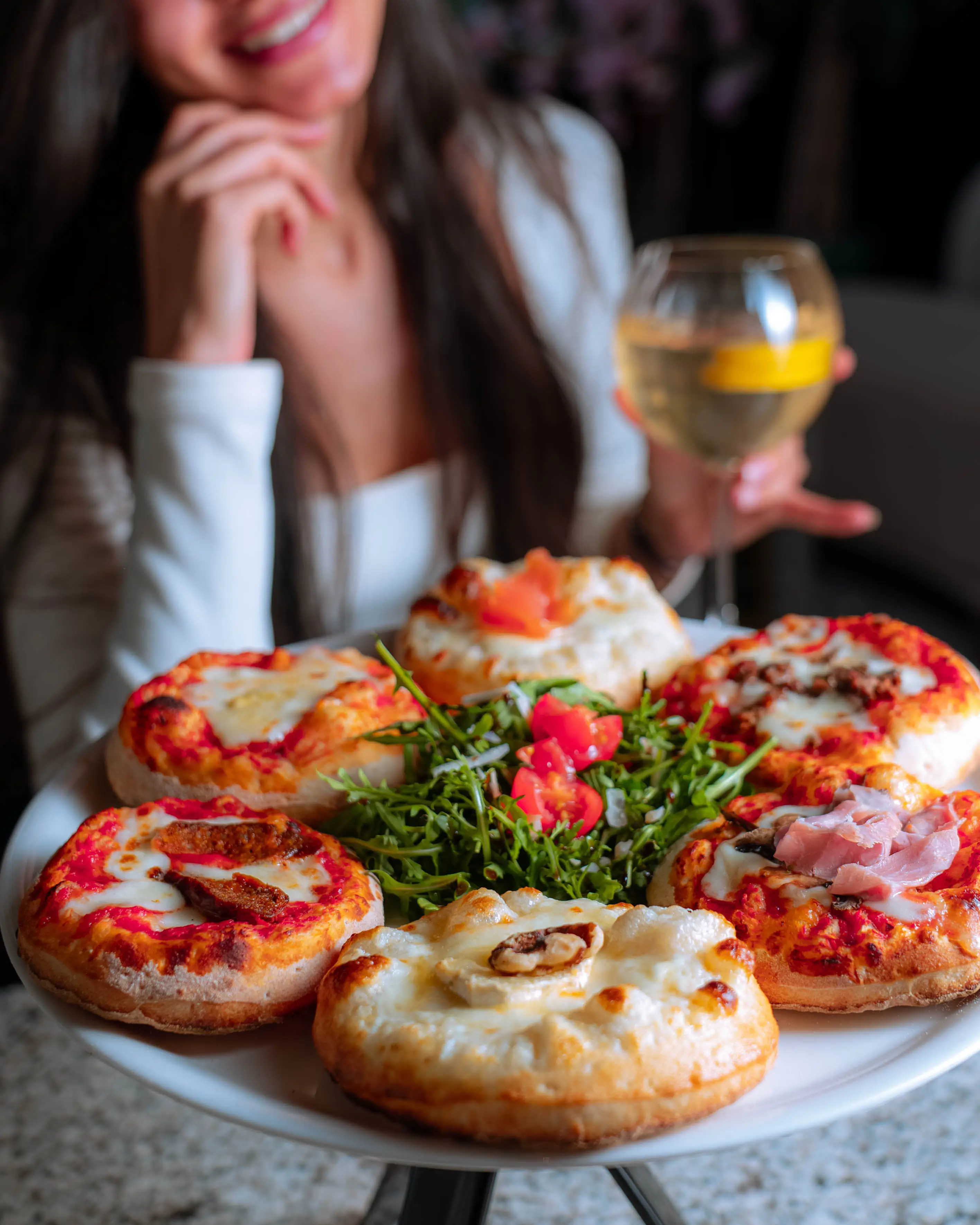Lebendige Restaurant-Szene: lachende Gästin mit einem Glas Weißwein vor einem Teller mini Pizzen.